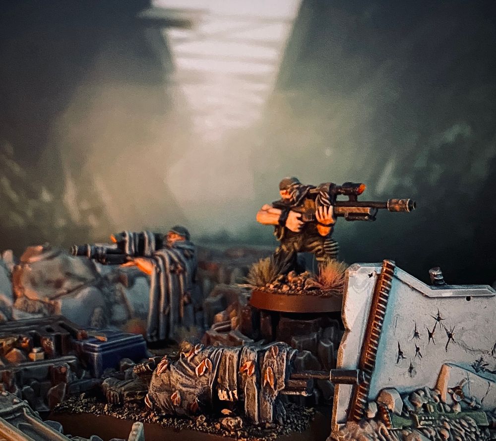 Three old Catachan Sniper miniatures (standing, kneeling and lying down) in different brown-green-grey tones set up in debris scatter terrain in front of a Backdrop showing a derelict hallway. 
The light from the left side looks like there’s a fire burning. 