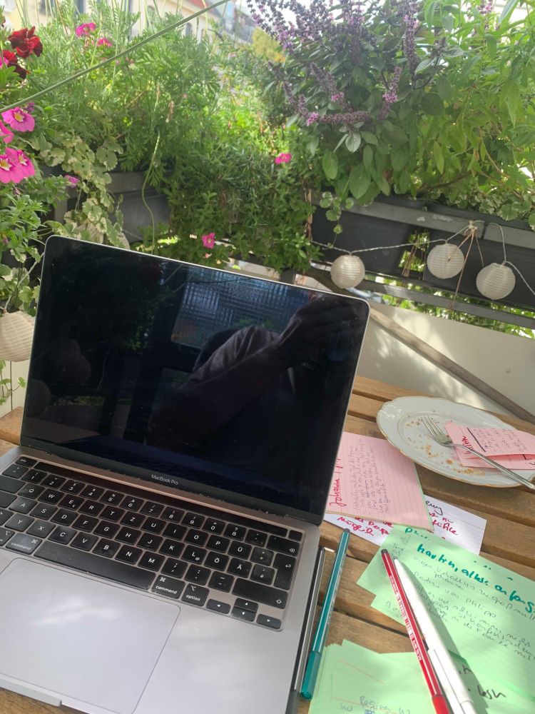 Foto von meinem Balkon. ein kleiner Tisch, auf dem ein aufgeklappter Laptop steht, auf dem Tisch liegen beschriebene Karteikarten und Stift. Am Geländer des Balkons stehen diverse Pflanzen und Blumen, es ist alles sehr grün und blüht.