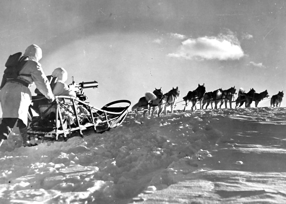 Two soldiers wearing winter camouflage sit in and push a dog sled with a machine gun mounted on it, behind a team of sled dogs. (U.S. National Archives)