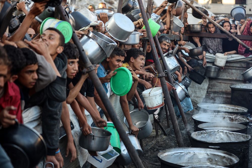 A large crowd of people stand along a fence, holding pots and plates at a food distribution center. (Majdi Fathi / NurPhoto / Getty)