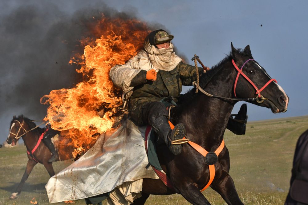 A rider on a horse, wearing flame-protective gear, rides with flames burning from their back, in a field. (Vladimir Voronin / AP)