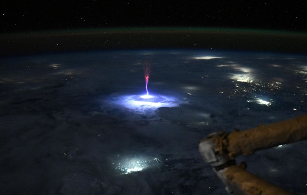 A nighttime view of the Earth, from orbit, with a bright blu-and-red bolt of electrical energy shooting up from a lightning strike in a storm below (Nichole Ayers / NASA)