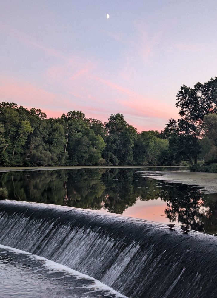 A tiny half-moon, seen in the sky above a small tree-lined reservoir and small dam.