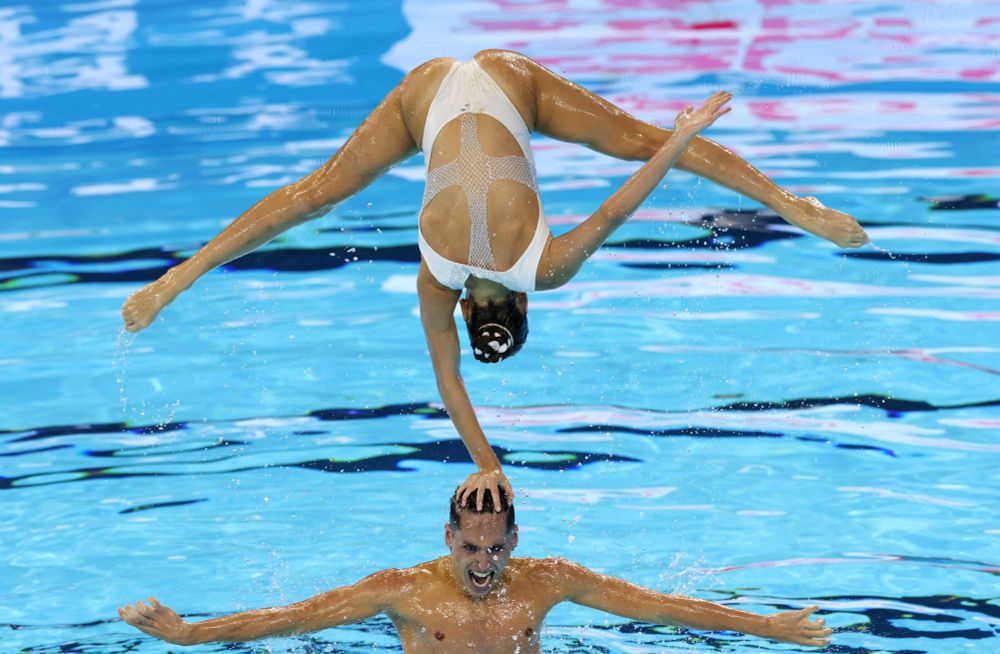 A pair of artistic swimmers seen during a performance, one of them, balancing on one hand placed on the head of the other, who is swimming in a pool. (Maye-E Wong / Reuters)