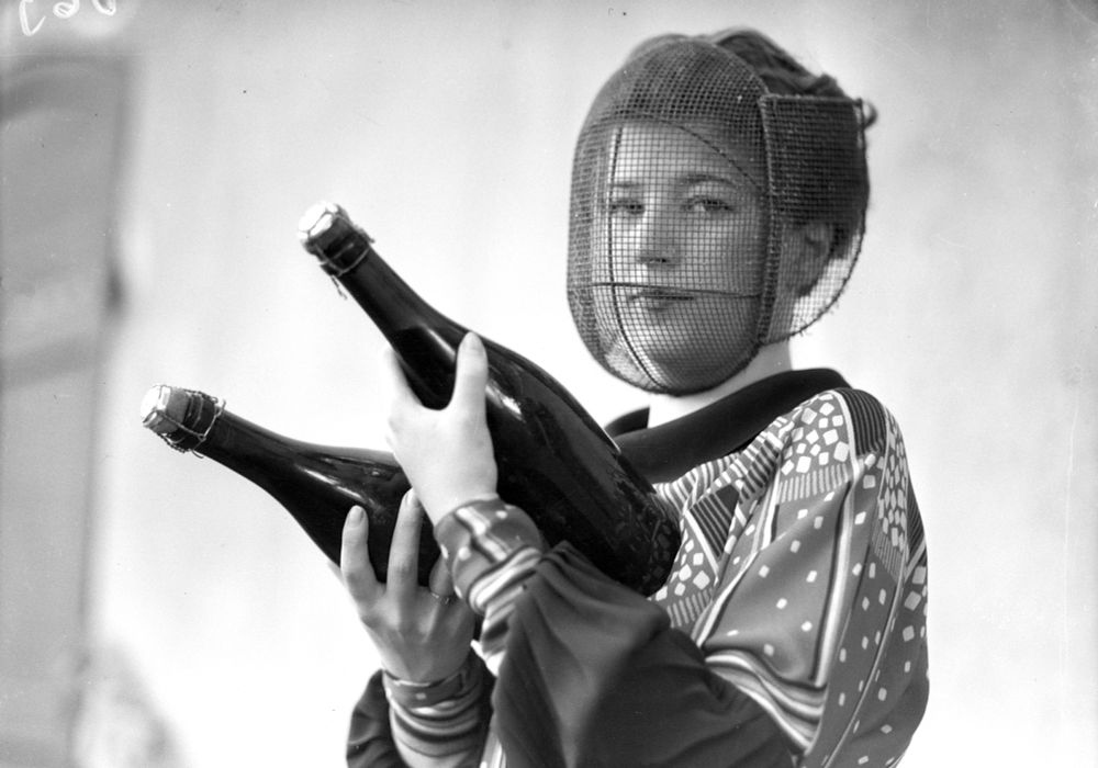 A person, a "champagne inspector," holds two champagne bottles in their hands, while wearing a protective mesh face mask. (Bettmann / Getty)