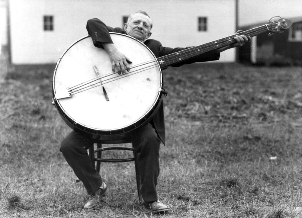 A person sitting on a stool outside plays a gigantic banjo. (Soibelman Syndicate / Visual Studies Workshop / Getty)