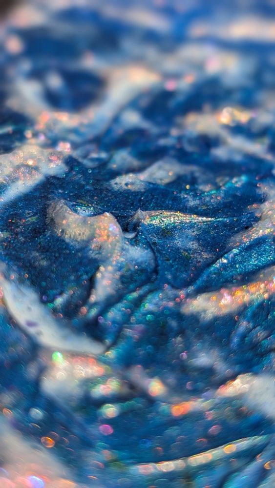 Macro detail view of a small metallic blue acrylic painting of abstract textured ocean waves. The crests of the waves are encrusted with enchanting opalescent sea foam exhibiting a rainbow sparkle and fiery pearlescence.