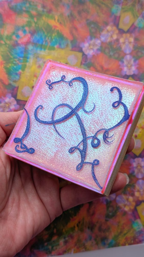 Close up of a hand holding a tiny colorful square acrylic painting over a colorful patterned background. The painting has a reflective neon pink background with a blue green iridescence. There are sparkly dark purple abstract swirls dancing over the background. 