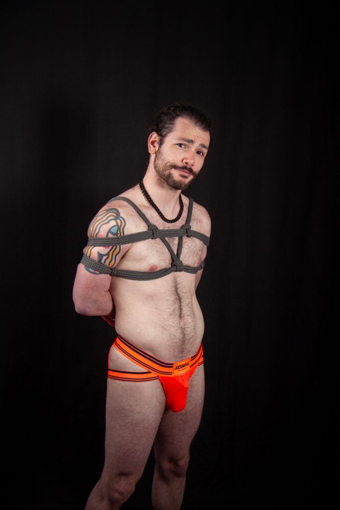 Bauhaus in a gray rope harness and orange briefs smirking at the camera