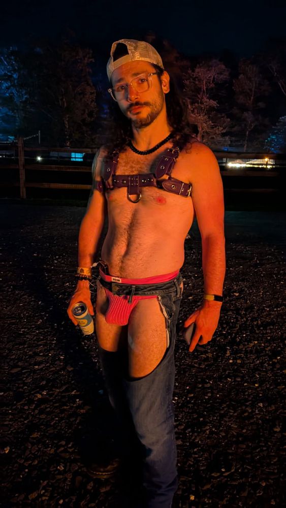 Bauhaus standing in a field at night wearing a harness, pink jockstrap, and denim chaps