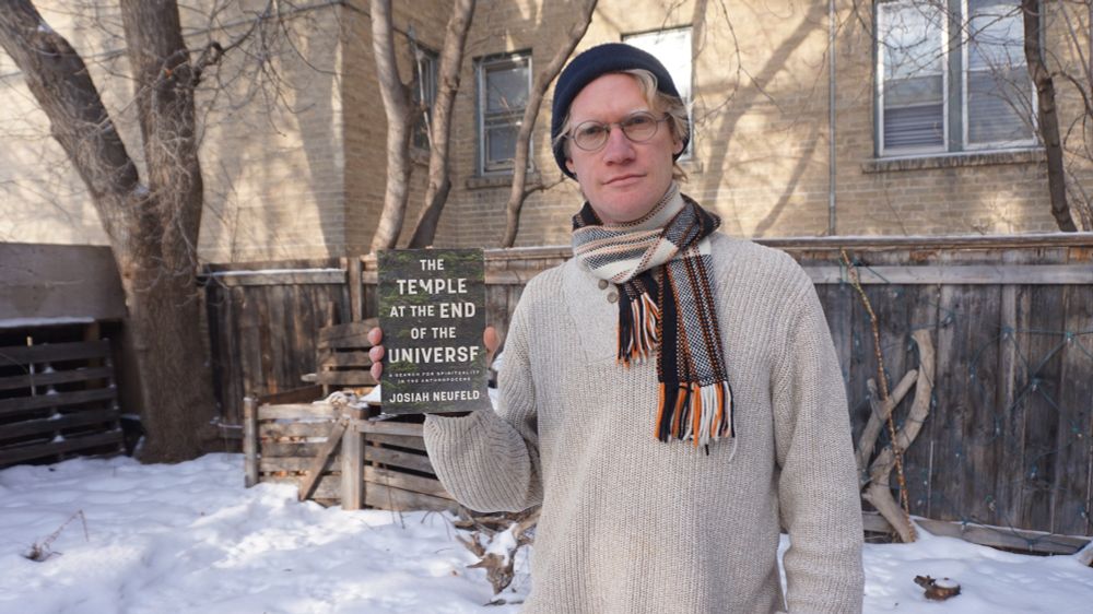 A person in a sweater and scarf standing outside in the snow holding a book titled The Temple at the End of the Universe.
