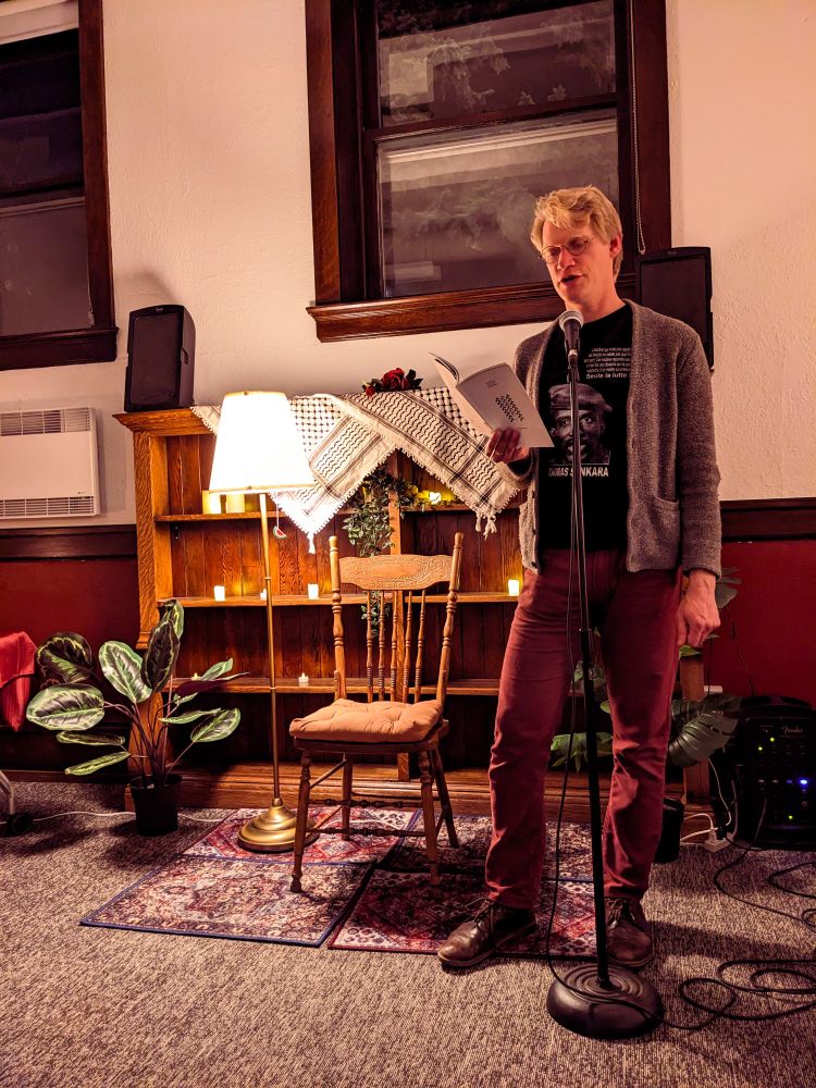 Person in a grey cardigan, black t-shirt and maroon pants reading from a book of poetry by Lee Maracle.