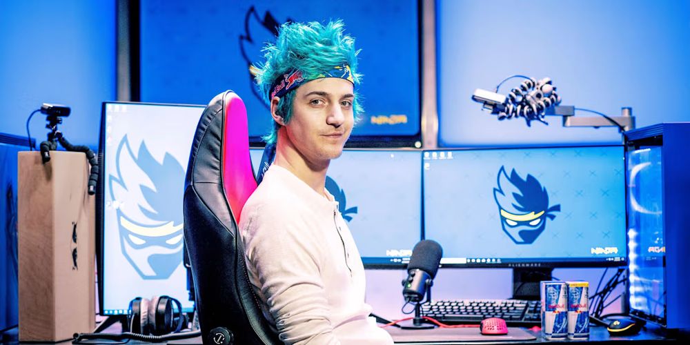 This is a picture of the streamer, gamer, and youtuber, Richard Tyler Blevins, otherwise known as Ninja. He is best known for Fortnite. He is sitting in his gaming chair looking at the camera, with a headband on and blue dyed hair. Behind him are multiple monitors and a full streaming setup, with the Ninja logo being shown on each monitor. One piece of his setup is also that he has two red bull cans on his desk net to his mouse.