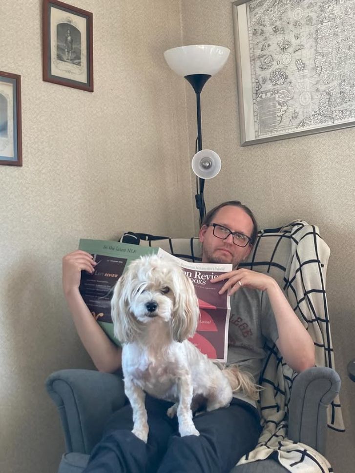 Alexander, a middle-aged man in glasses, sits in an armchair reading a magazine. Laban, a white bichon dog with his fur cut short, sits in his lap. Laban is looking into the camera. He's got slightly longer hair on top of his head, and some strands have fallen down over his right eye.