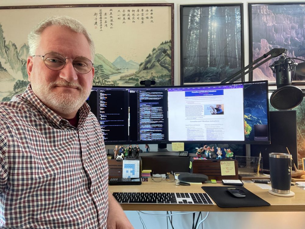 photo of Dan Russell (white male, approximately 55 years old, gray hair, beard, and glasses, wearing a checkered shirt) standing to the left of a computer monitor.