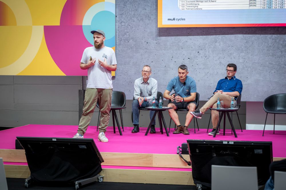 The accessible ALT text in English follows below

Deutsch – Vortrag auf der Cargo Academy der Eurobike 2025: Der Head of Marketing von muli-cycles steht auf der Bühne und spricht über werkstattspezifische Prozesse und Servicequalität im Cargobike-Sektor. Thema ist unter anderem die Arbeitswerteliste als Grundlage für transparente Abrechnung und effiziente Reparaturprozesse. Auf dem Podium sitzen Vertreter weiterer Hersteller und des Branchenverbands Zukunft Fahrrad. Im Hintergrund ist das muli Logo auf der Präsentationsfolie zu sehen.

English – Presentation at the Cargo Academy at Eurobike 2025: The Head of Marketing from muli-cycles speaks on stage about workshop processes and service quality in the cargo bike industry. Key topic: the work value sheet as a reference for transparent invoicing and efficient repairs. On the panel sit other manufacturers and a representative from the industry association Zukunft Fahrrad. The muli logo is visible on the presentation slide in the background.