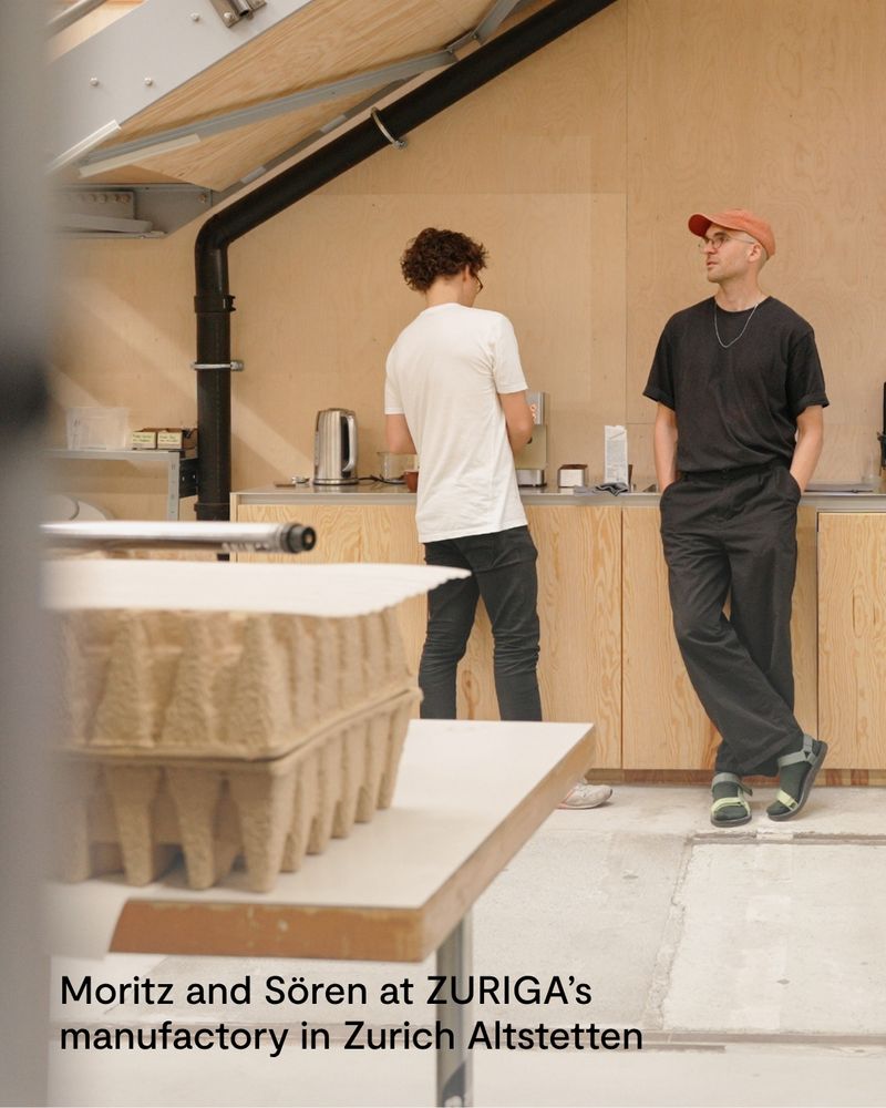 The accessible ALT text in English follows below

Deutsch – Foto von Moritz Güttinger und Sören Gerhardt in der ZURIGA-Manufaktur in Zürich Altstetten. Beide stehen in einem hellen Holzraum an einer Arbeitsfläche mit Espressomaschine und Wasserkocher. Im Vordergrund sind Verpackungstrays aus Pappe zu sehen.

English – Photo of Moritz Güttinger and Sören Gerhardt at the ZURIGA manufactory in Zurich Altstetten. They are standing in a light wooden space at a counter with an espresso machine and kettle. Cardboard trays appear in the foreground.
