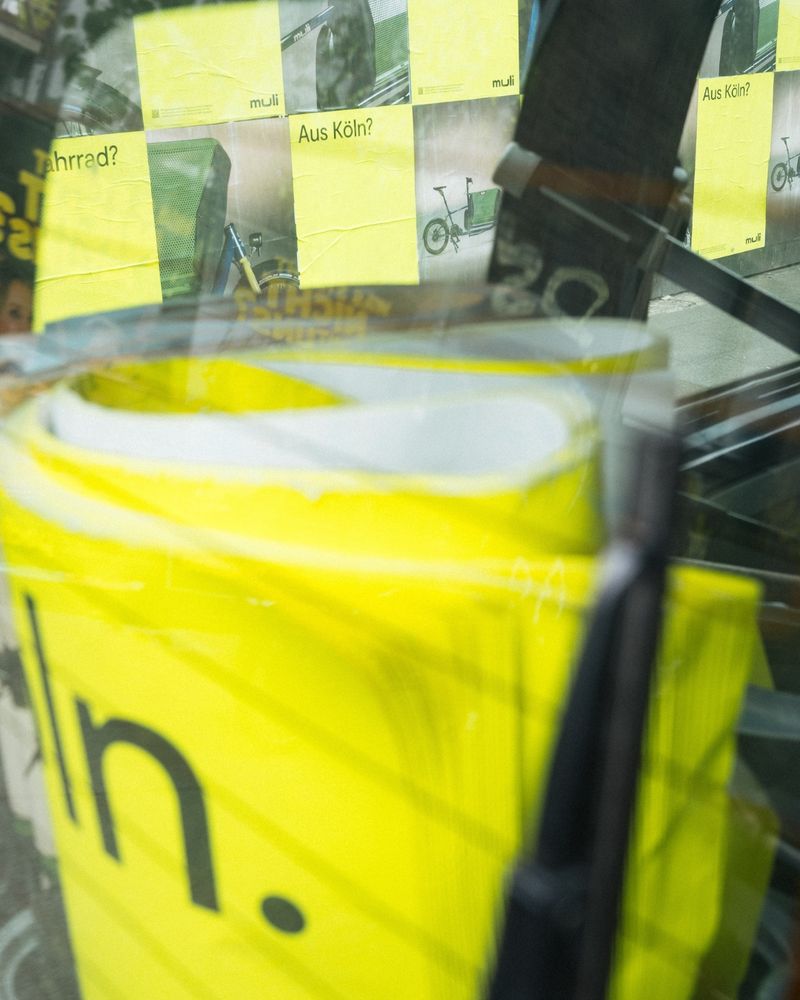 The accessible ALT text in English follows below

Deutsch – Durch eine Autoscheibe fotografierte Szene mit gerollten und geklebten muli-Plakaten in leuchtendem Gelb. Aufschriften wie „Ein Fahrrad?“ und „Aus Köln?“ machen auf die Herkunft der muli Lastenräder aufmerksam. Teil der ersten offiziellen Plakatkampagne von muli in Köln mit 4.000 Plakaten im gesamten Stadtgebiet.

English – Photo taken through a car window showing rolled-up and pasted bright yellow muli posters with texts like “A bike?” and “From Cologne?”. Part of muli’s first official poster campaign with 4,000 posters across the city, highlighting the local origin of the cargo bikes.