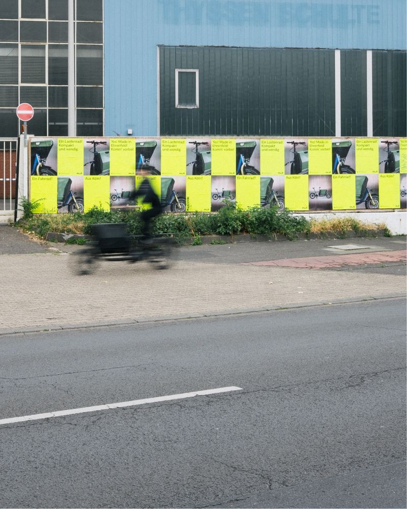 The accessible ALT text in English follows below

Deutsch – Plakatwand an einem Industriegebäude in Köln, beklebt mit auffälligen muli-Postern. Aussagen wie „Ein Lastenrad? Kompakt und wendig.“ und „Yes! Made in Ehrenfeld!“ präsentieren muli als lokal produziertes, innovatives Cargobike. Im Vordergrund verschwommene Bewegung eines muli Cargobikes. Teil der ersten großen muli-Kampagne mit 4.000 Plakaten in Köln.

English – Poster wall at an industrial building in Cologne covered with striking muli posters. Statements like “A cargo bike? Compact and agile.” and “Yes! Made in Ehrenfeld!” promote muli as a locally produced, innovative cargo bike. Blurred motion of a muli cargo bike in the foreground. Part of muli’s first major campaign with 4,000 posters across Cologne.