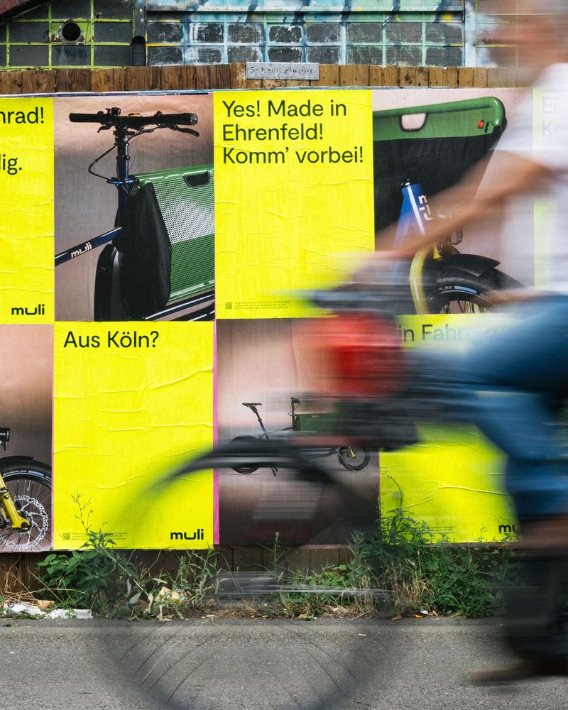 The accessible ALT text in English follows below

Deutsch – Nahaufnahme von muli-Plakaten an einer Kölner Wand mit Graffiti. Texte wie „Yes! Made in Ehrenfeld!“ und „Ein Fahrrad?“ rücken die lokale Produktion des muli in den Fokus. Eine Person auf einem roten Fahrrad fährt dynamisch durchs Bild. Teil der ersten großflächigen muli-Plakatkampagne mit 4.000 Plakaten in Köln.

English – Close-up of muli posters on a graffiti-covered wall in Cologne. Slogans like “Yes! Made in Ehrenfeld!” and “A bike?” emphasize the local production of the muli. A person on a red bicycle passes dynamically through the frame. Part of muli’s first large-scale poster campaign with 4,000 posters in Cologne.