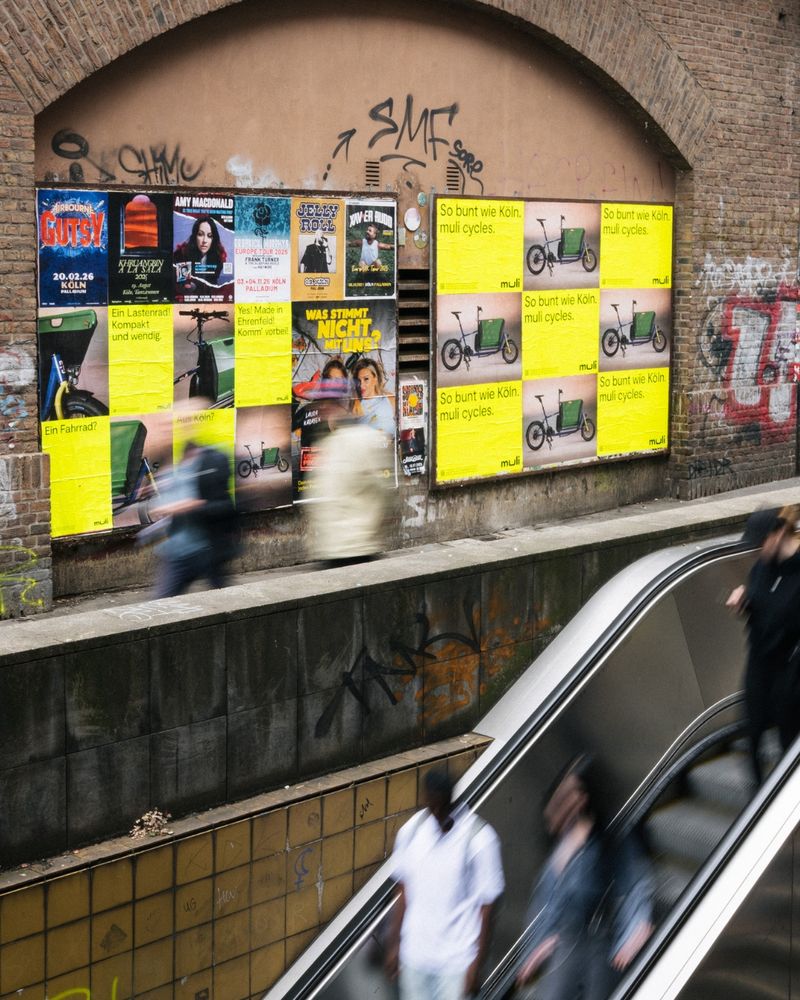 The accessible ALT text in English follows below

Deutsch – U-Bahn-Ausgang mit Rolltreppe in Köln. An der Wand: eine Vielzahl von muli-Plakaten mit der Botschaft „So bunt wie Köln. muli cycles.“ Im urbanen Umfeld bewegen sich Menschen durch das Bild. Die Szene ist Teil der ersten offiziellen muli-Kampagne mit 4.000 Plakaten, die in ganz Köln platziert wurde – laut, sichtbar und mit klarer Haltung.

English – Subway exit with an escalator in Cologne. On the wall: numerous muli posters with the message “As colorful as Cologne. muli cycles.” People move through the urban setting. The scene is part of muli’s first official campaign with 4,000 posters placed all over Cologne – loud, visible, and with a clear message.