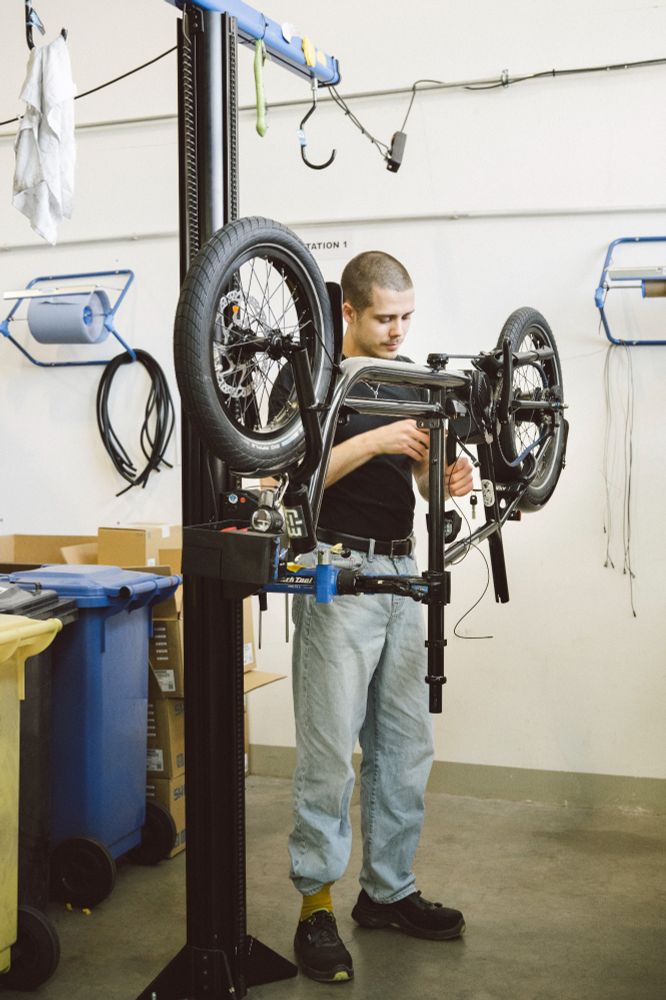 Kevin from the muli assembly team is working on a muli ST. The bike is mounted upside down on a stand in a clean, well-organized workshop in Cologne. Kevin is focused on wiring or adjusting parts near the frame, surrounded by tools and components.