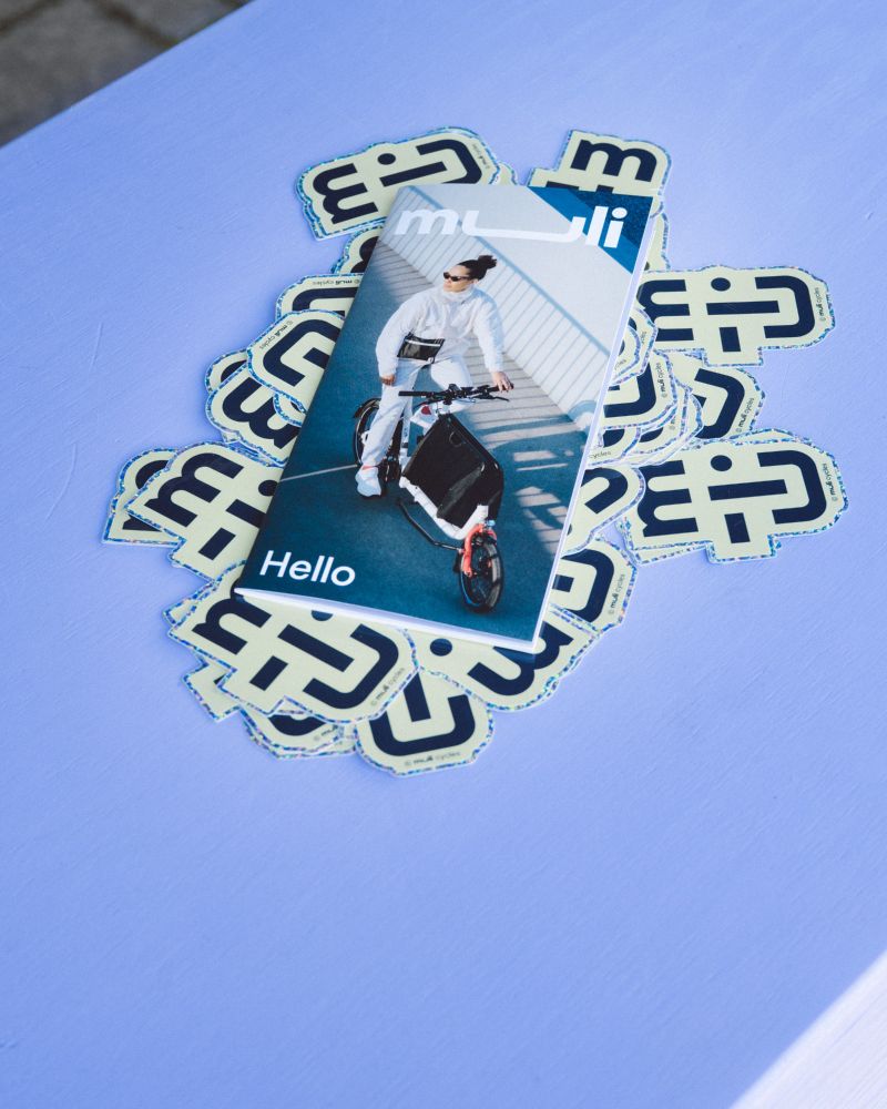 text in English follows below

Deutsch – Auf einem lilafarbenen Tisch liegen Sticker mit dem muli Logo sowie ein Faltflyer mit der Aufschrift „Hello“. Auf dem Cover des Flyers ist eine Person mit weißem Outfit auf einem muli Bike abgebildet.

English – On a lavender-colored table are stickers with the muli logo and a folded flyer titled “Hello.” The cover of the flyer shows a person in a white outfit on a muli bike.
