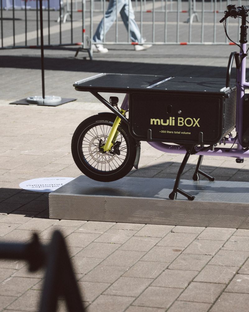 text in English follows below

Deutsch – Nahaufnahme der muli BOX auf einem lilafarbenen Ausstellungsbike auf der Eurobike. Die Box ist verschlossen und trägt den Aufdruck „muli BOX ~350 liters total volume“. Eine Person geht im Hintergrund vorbei.

English – Close-up of the muli BOX on a purple demo bike at Eurobike. The box is closed and labeled “muli BOX ~350 liters total volume.” A person is walking in the background.