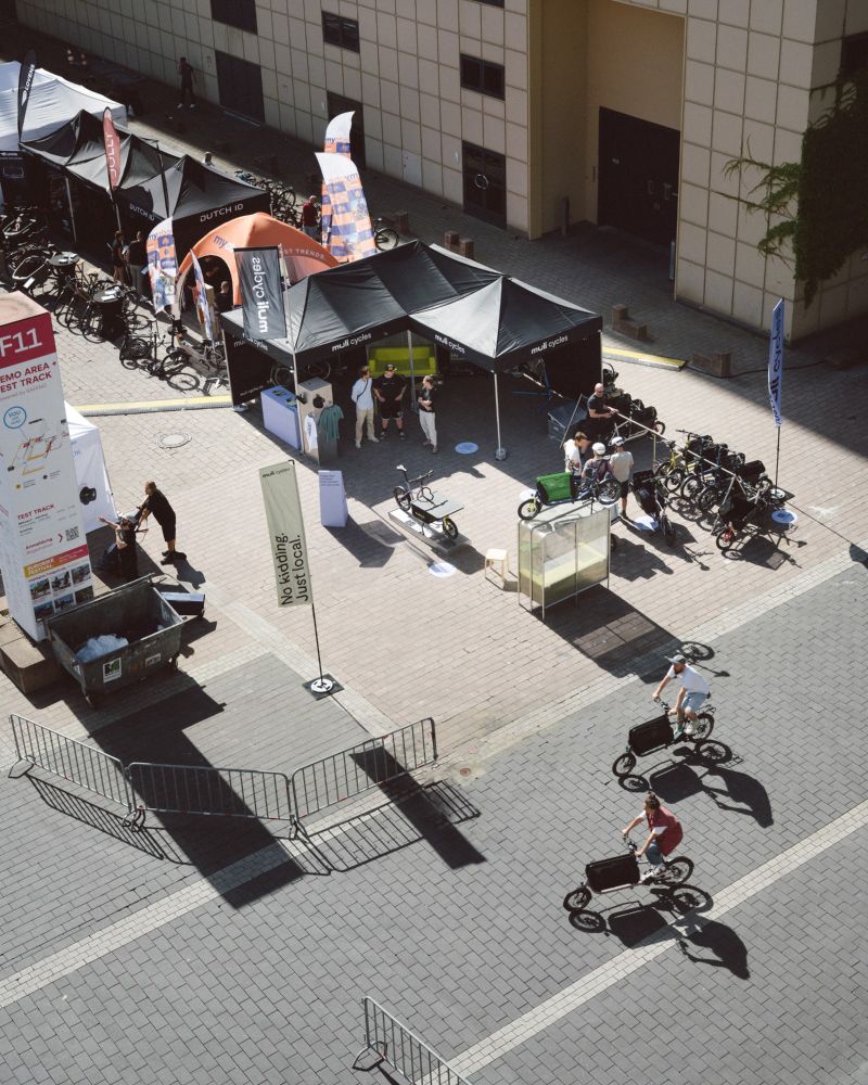 The accessible ALT text in English follows below

Deutsch – Blick von oben auf den muli-cycles Messestand bei der Eurobike. Schwarze Zelte mit muli Branding, Testbikes und Besucher:innen sind zu sehen. Zwei Personen fahren auf muli Rädern durch den Testbereich.

English – Top-down view of the muli-cycles booth at Eurobike. Black tents with muli branding, test bikes, and visitors are visible. Two people are riding muli bikes through the test area.