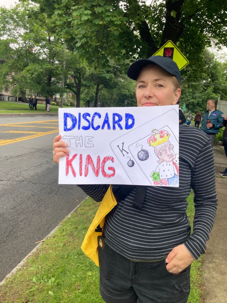 Woman holding a sign with a picture of a playing card that says “Discard The King” 