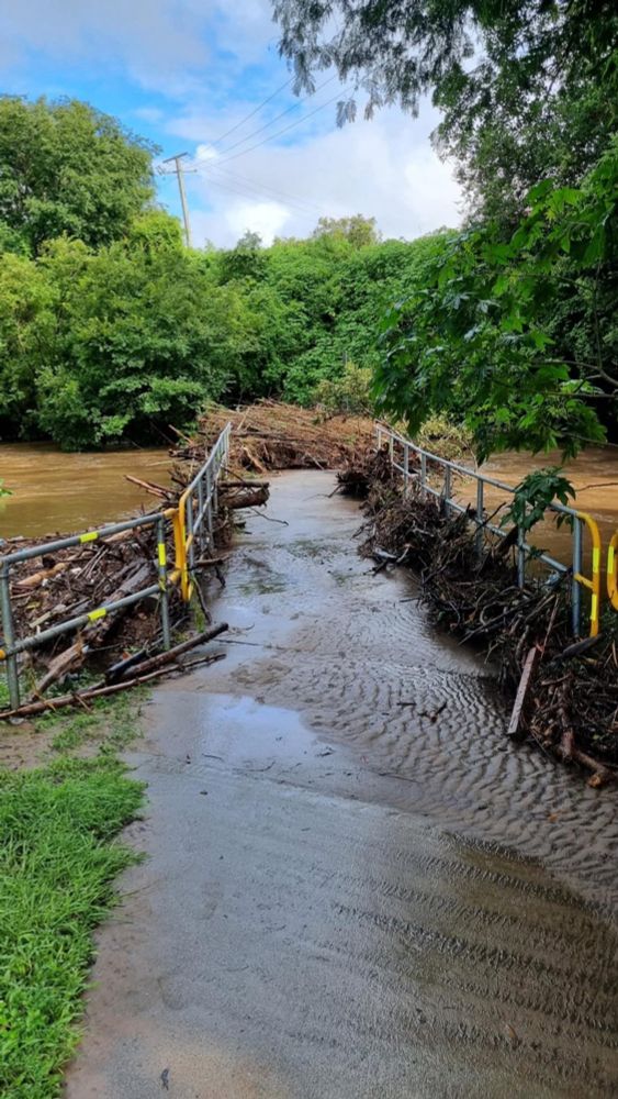 A flooded path leading to a pedestrian bridge covered with debris and a flooded brown river running over the bridge. 