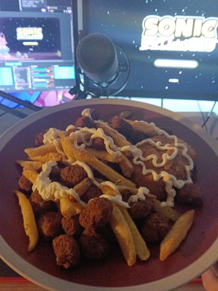 A plate of chips, popcorn chicken, and a chicken kiev covered in mayo in front of a desktop computer 