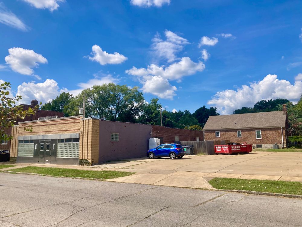 One story circa 1940 commercial brick building with a large parking lot on the side. A dumpster and port-a-potty are on the lot. A brick residence is behind the lot.