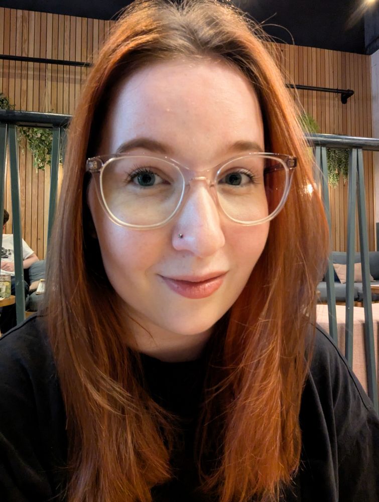 Selfie of me smiling. I have brunette hair with red balayage, clear framed glasses, simple makeup, a (new) nose piercing, and a black tshirt on.