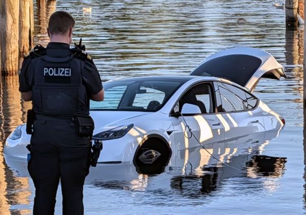 ein Polizist steht mit dem Rücken zur Kamera und schaut auf einen weißen Tesla, der mit offener Kofferraumklappe zu tief im Wasser steht