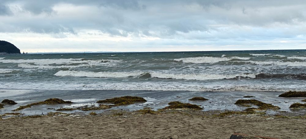 Wellen mit Gischt brechen sich, als sie auf den Strand zu rollen
es ist ein Tag mit Wind und Wetter, man sieht die Wolken quer über den Himmel in unterschiedlichen Höhen und Formen, darunter die aufgewühlte See
