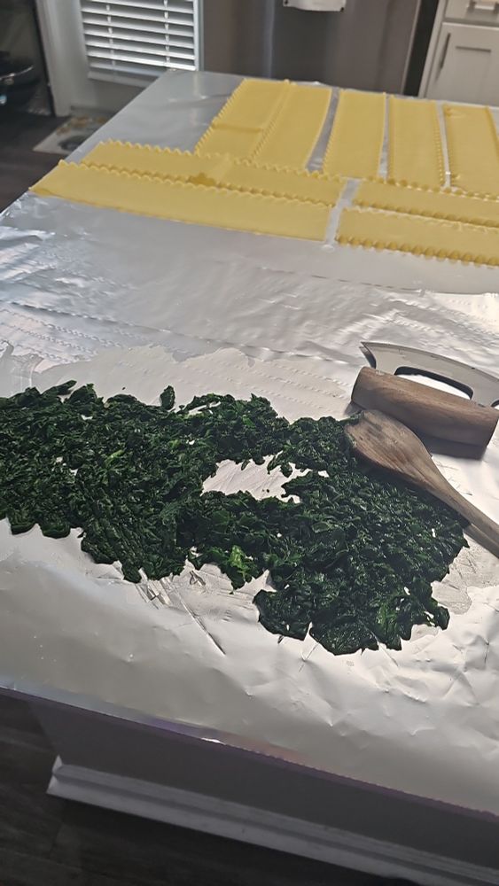 Fresh spinach, sauteed down and chopped, laid out on foil. Lasagna noodles laid out on foil behind it. Wooden spoon and Ulu knife next to spinach. 