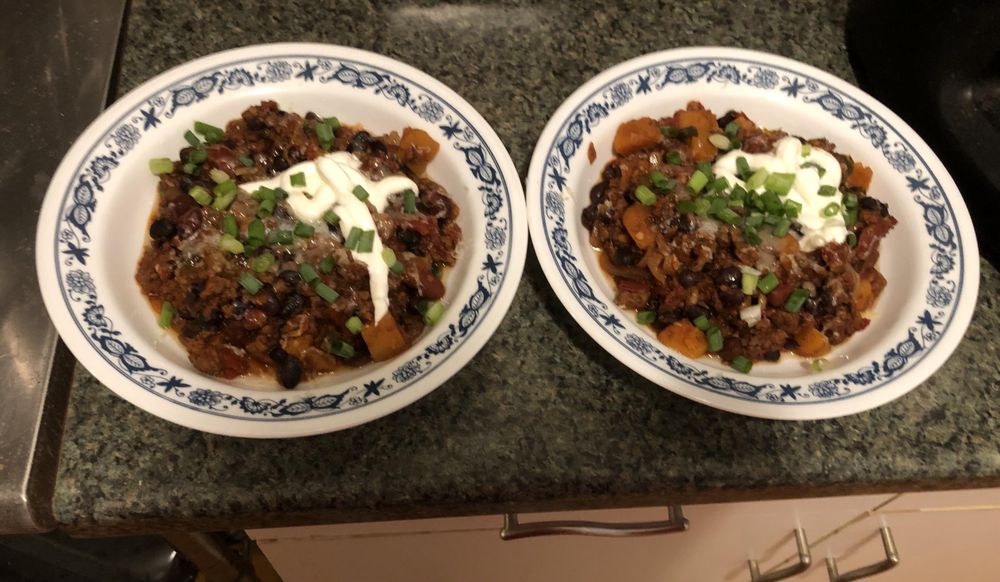 Chili plated in 2 flat bowls, topped with cheese, sour cream, scallions, and lime juice.