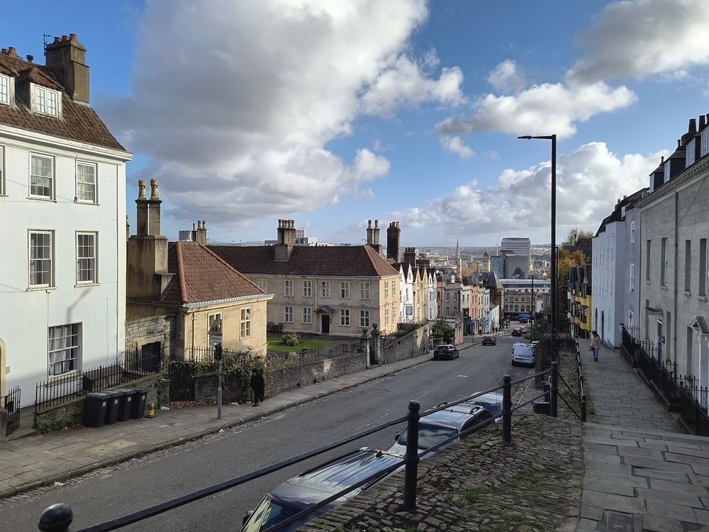 A view looking downhill on a very steep street in Bristol. Georgian houses on both sides of the road, with an almshouse in the foreground. The pavement is steeply raised up on one side of the road, and has frequent steps
