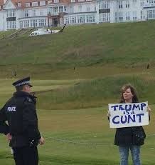 Janey Godley standing in front of a police officer holding a placard which reads 'Trump is a cunt'.
