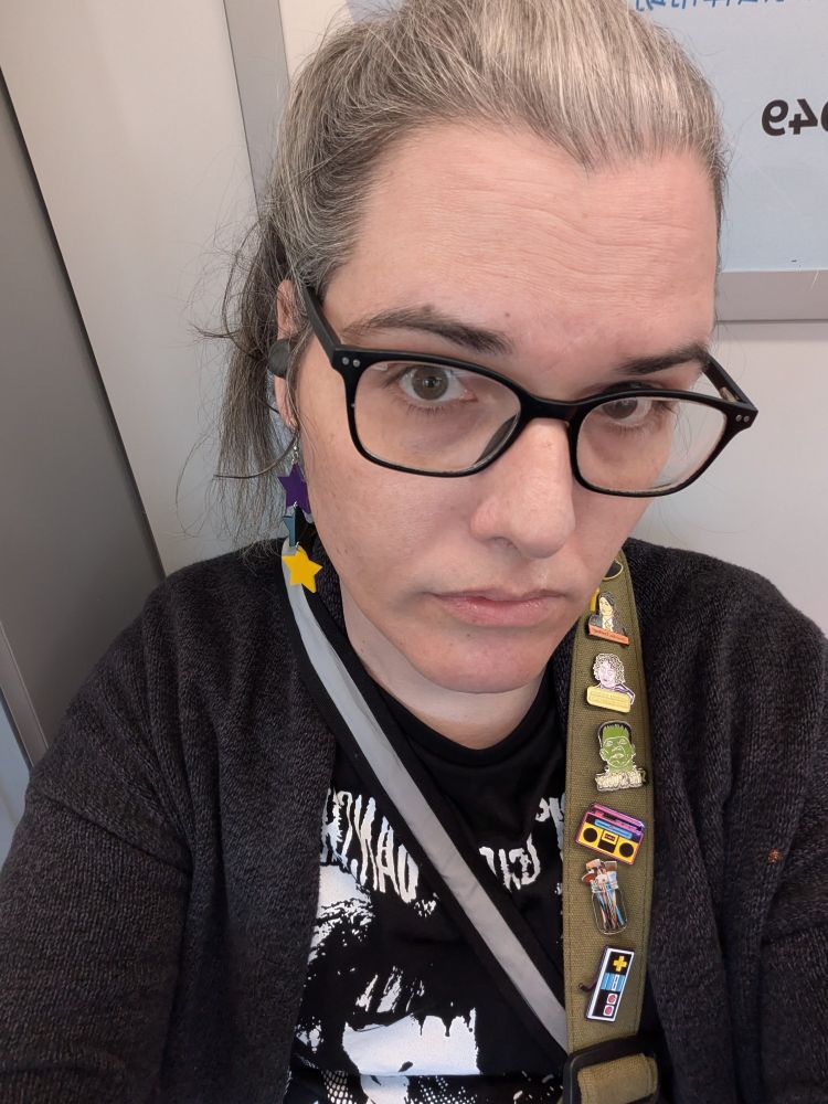 A selfie of me, a woman-passing enby wearing my hair in a pony tail & with dangle purple, black & yellow star earrings