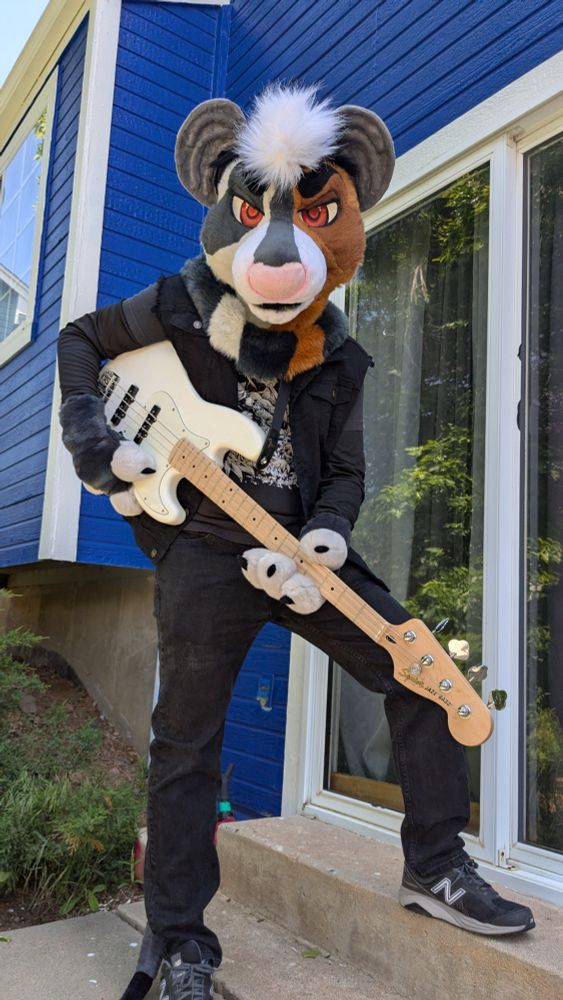 A person wearing a rat partial. He sports a black jean vest, a metal looking shirt, and is holding a bass guitar as if playing it.
