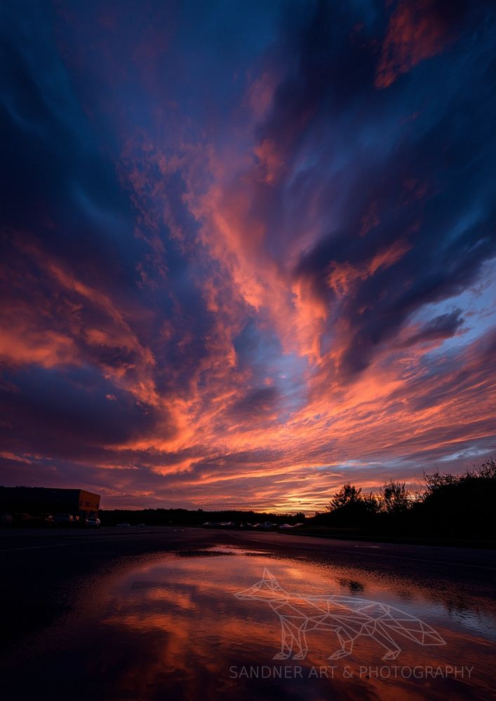 A dramatic sunset fills the sky with vibrant orange, red, and dark blue clouds, which are reflected in a large puddle on a wet asphalt surface in the foreground. Silhouetted trees and what appears to be a building are visible on the horizon.