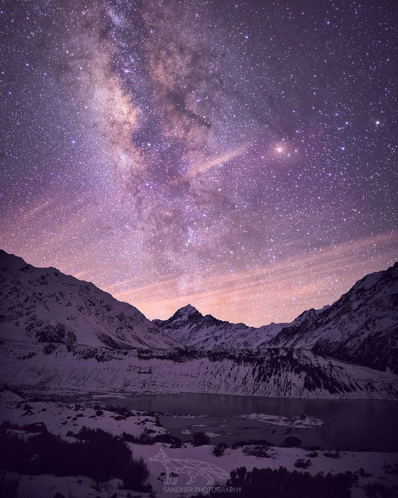 A dazzling view of the Milky Way stretches across the sky above snow-covered peaks in Aoraki / Mount Cook National Park. The galactic core glows brightly in hues of lavender, gold, and deep violet, illuminating thin wisps of interstellar dust. Below, rugged alpine mountains frame a frozen glacial lake, with scattered ice chunks floating quietly on its surface. The serene landscape is bathed in soft, starlit tones, merging the majesty of earth and cosmos into a single timeless scene.

