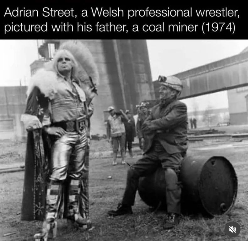 A picture of a coal miner, in his work gear, sitting on a 44 gallon drum. Standing beside him is a professional wrestler in make up, wearing high heels, and a cape, with long blond hair. He is looking to the sky past the camera.

The photo is captioned "Adrian Street, a Welsh professional wrestler, pictured with his father, a coal miner.