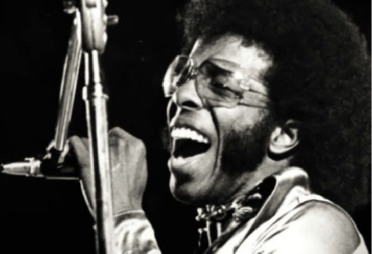 A black and white photo of Sly Stone singing into a microphone.