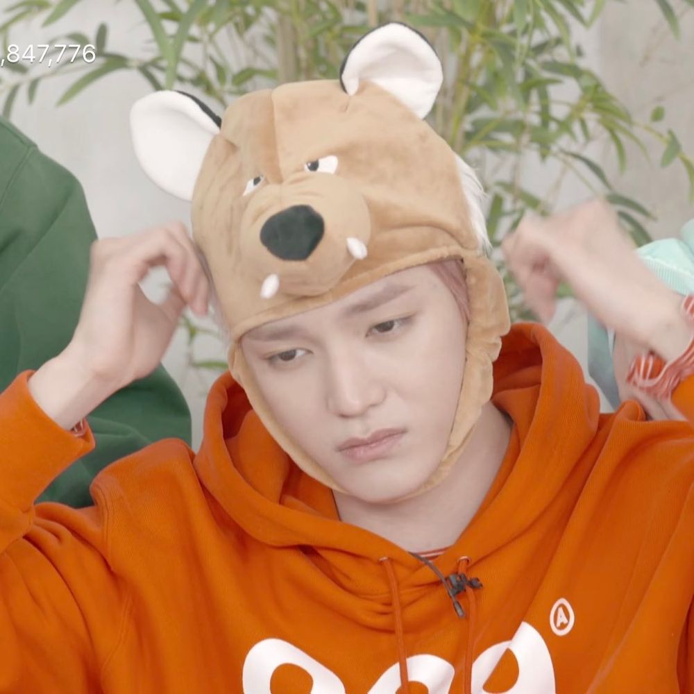 NCT 127's Taeyong wearing a bright orange hoodie and adjusting a silly wolf/bear hat on his head during a 2018(?) VLive