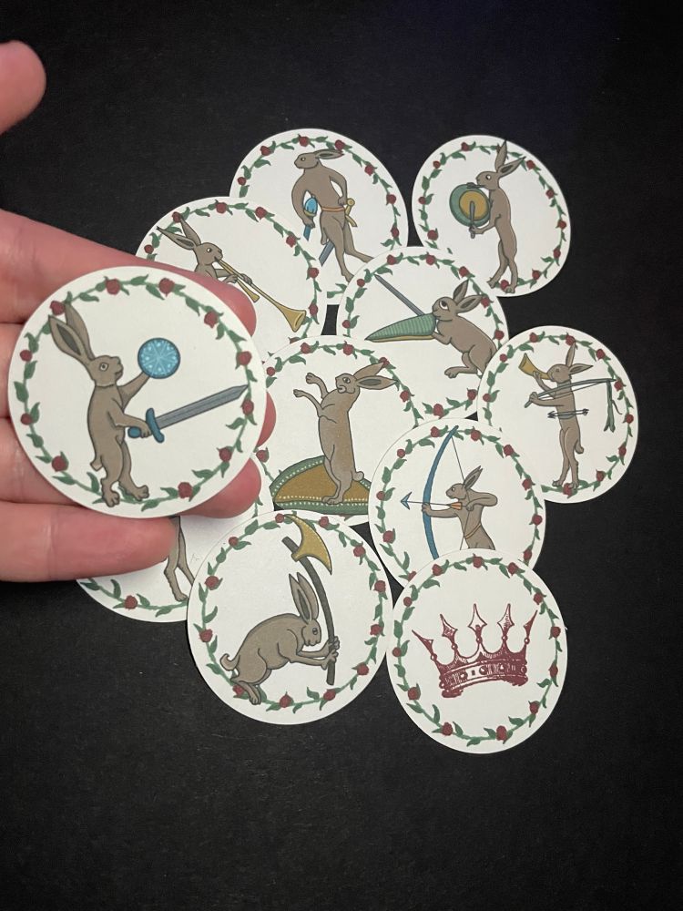Hand holds medieval warrior rabbit with sword and shield whole behind are a pile of similar medieval warrior and musical rabbits and one with red crown, all are surrounded by green floral border with red roses
