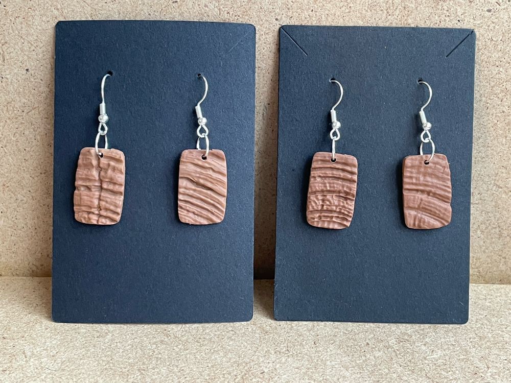 Two pairs of rectangular small earrings woth linear textured brown surface on black card on light brown background