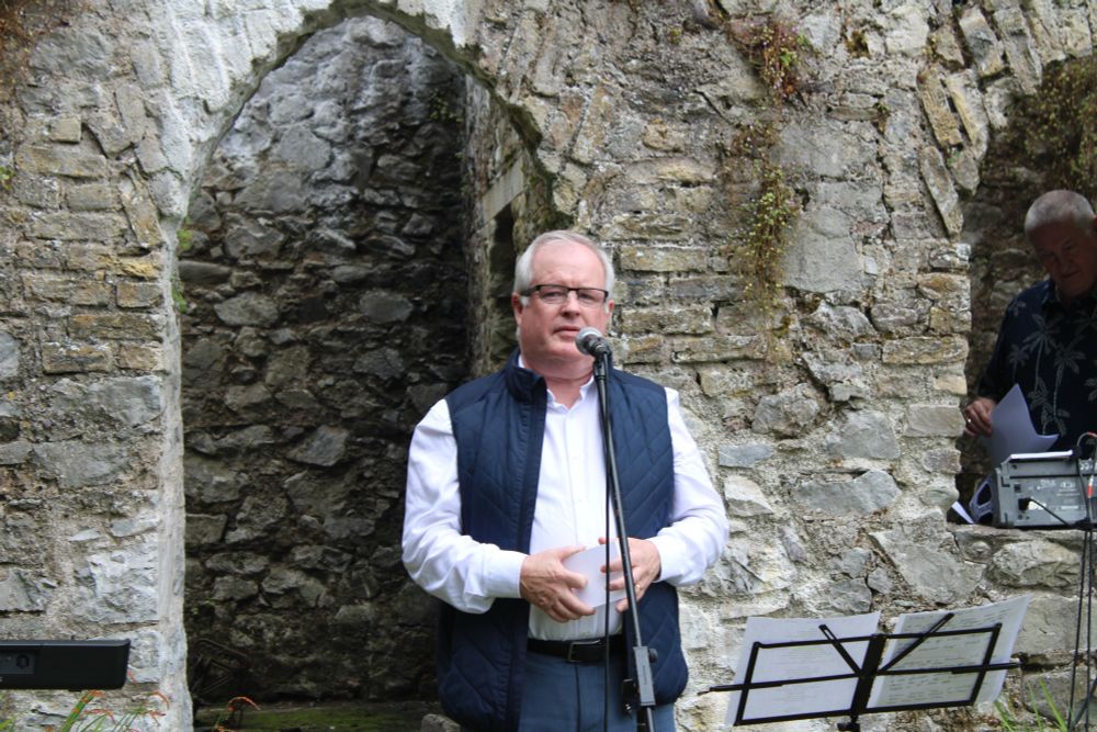 A man in a white shirt wearing a sleeveless jacket speaks into a microphone.